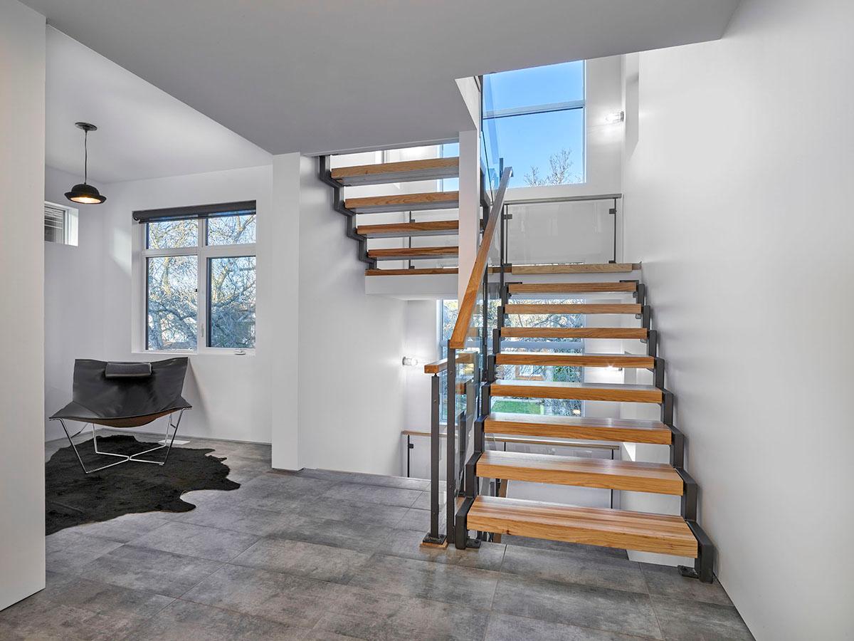 Modern floating staircase with wood treads, steel supports, and glass railings, adjacent to large windows and a contemporary seating area.