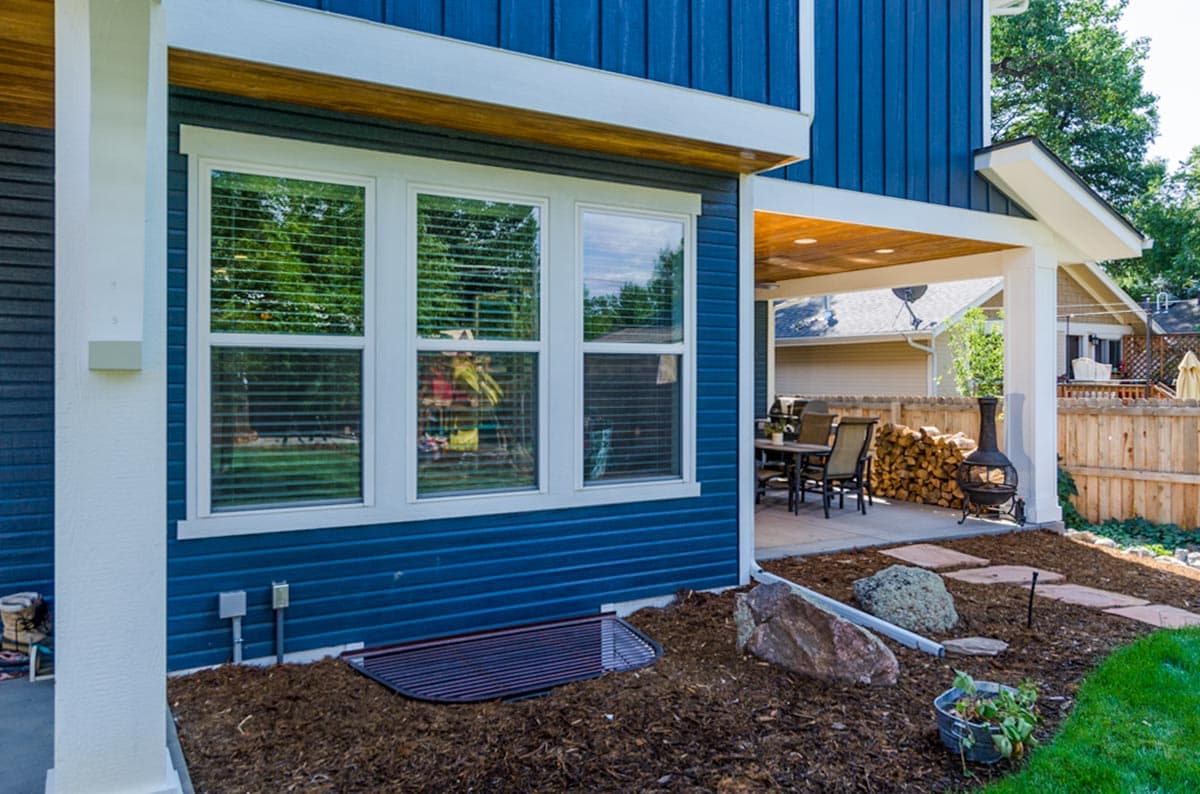 Exterior view of blue siding house with covered patio, three windows, and window well.