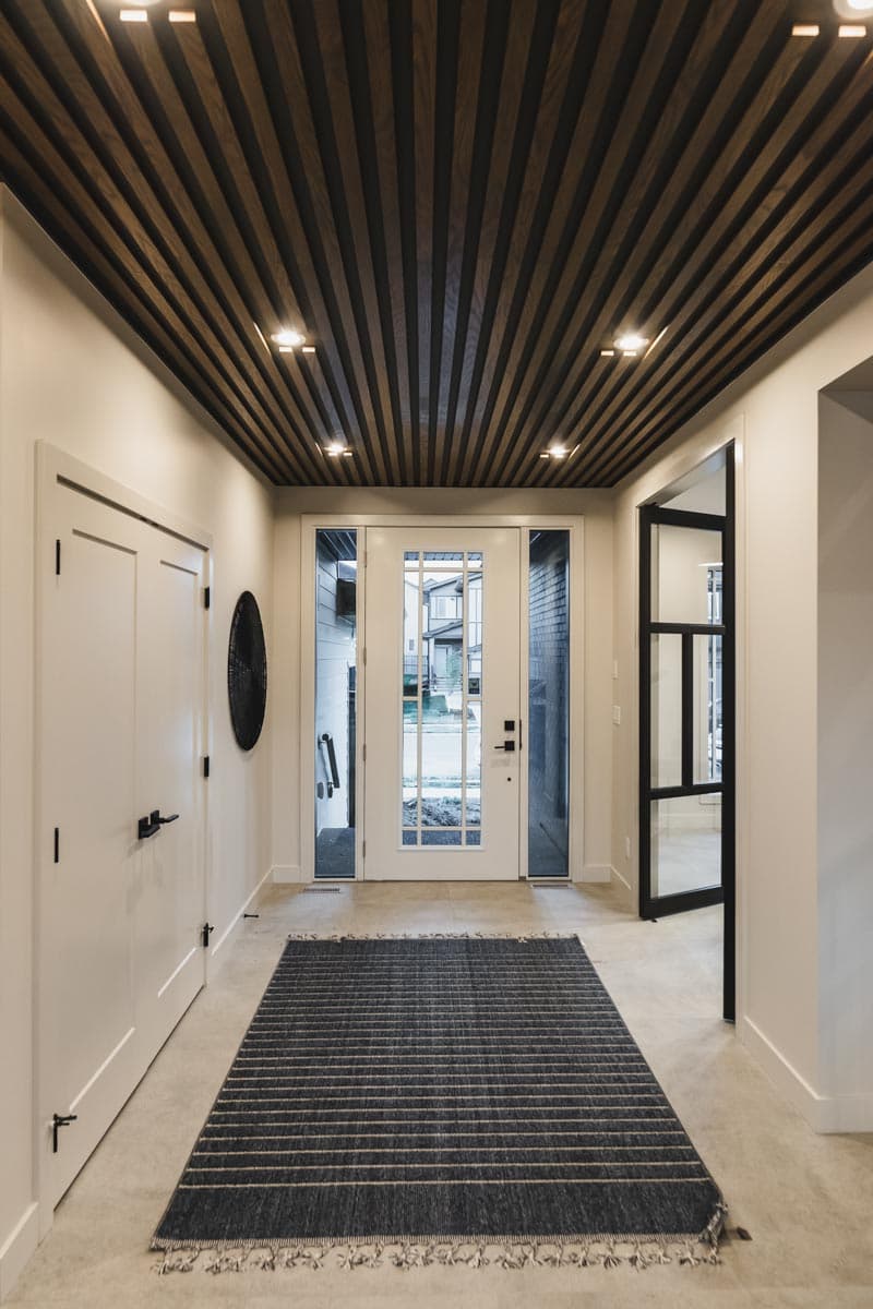 Modern entryway with slatted wood ceiling, double glass doors, and a striped rug on concrete flooring.