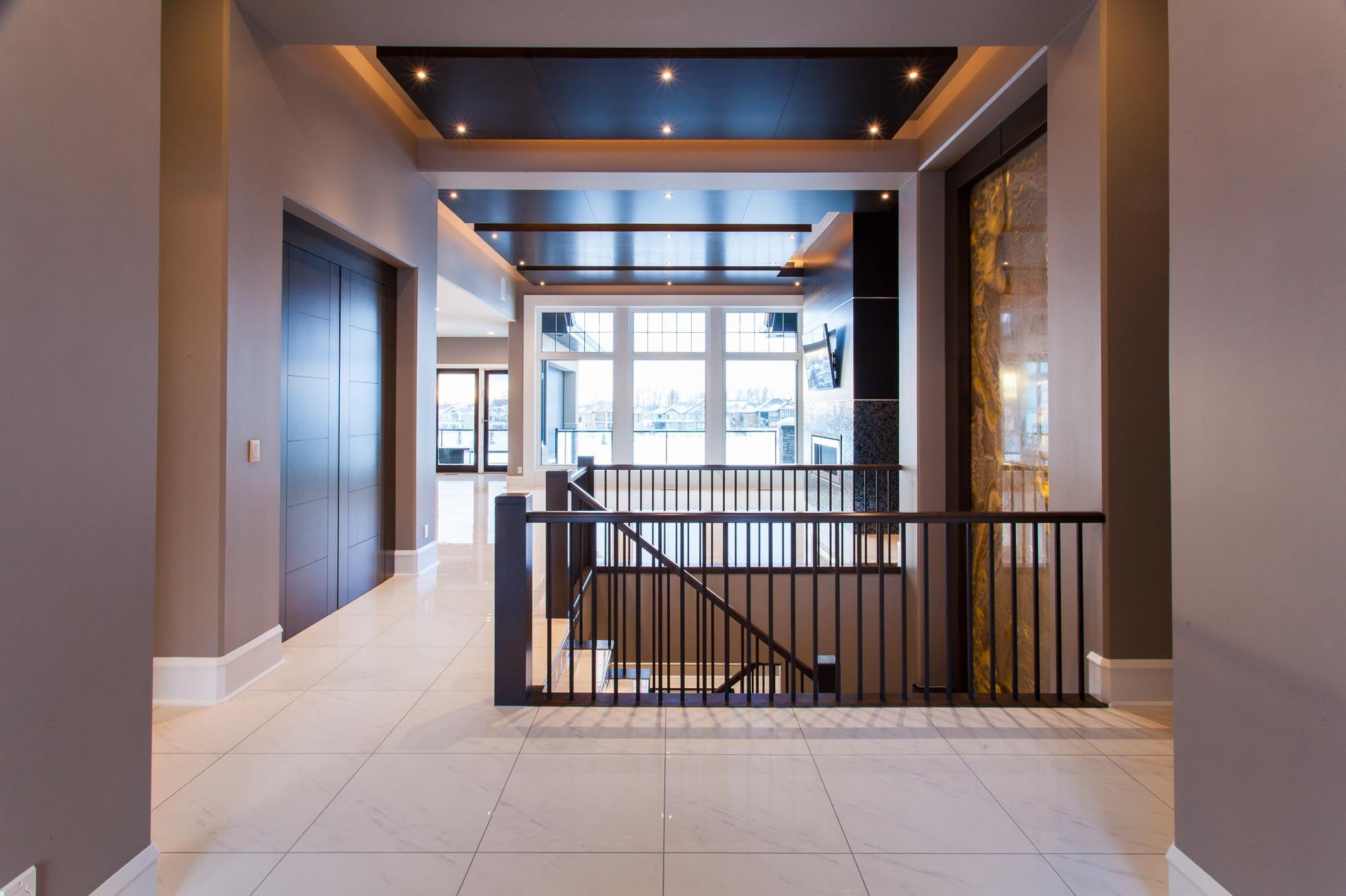 Interior view of a modern house hallway with a coffered ceiling, large windows, and a dark wood staircase railing.