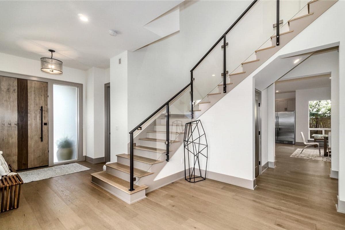 Modern entryway with wood plank doors, glass railing staircase, and view into kitchen with stainless steel appliances.