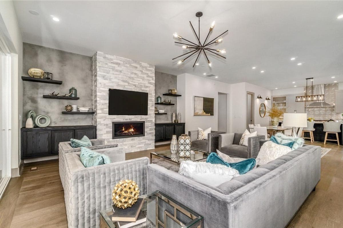 Living room with stone fireplace, built-in shelving, and modern chandelier. Open to kitchen area.