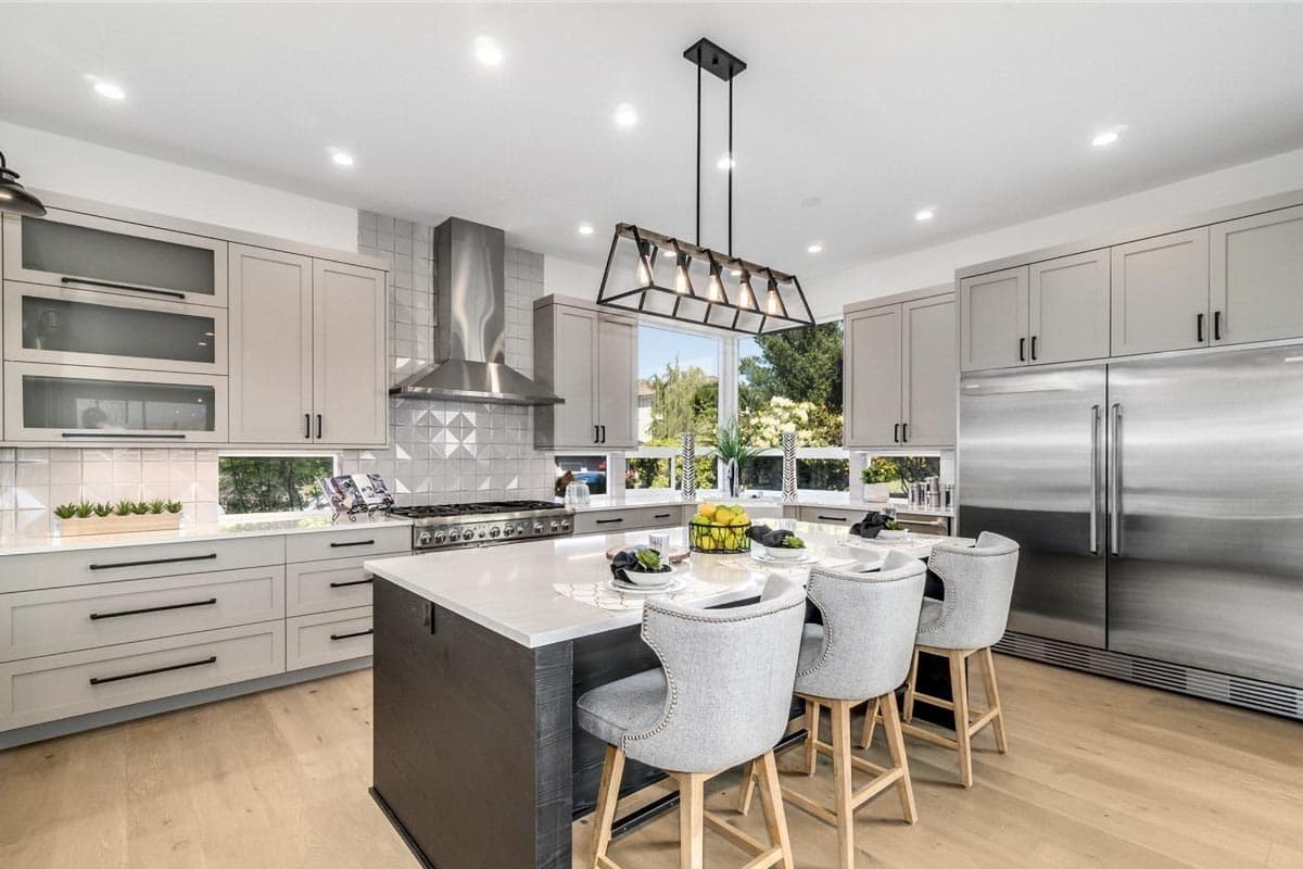 Contemporary kitchen with island, stainless steel appliances, range hood, light gray cabinetry, and large windows.