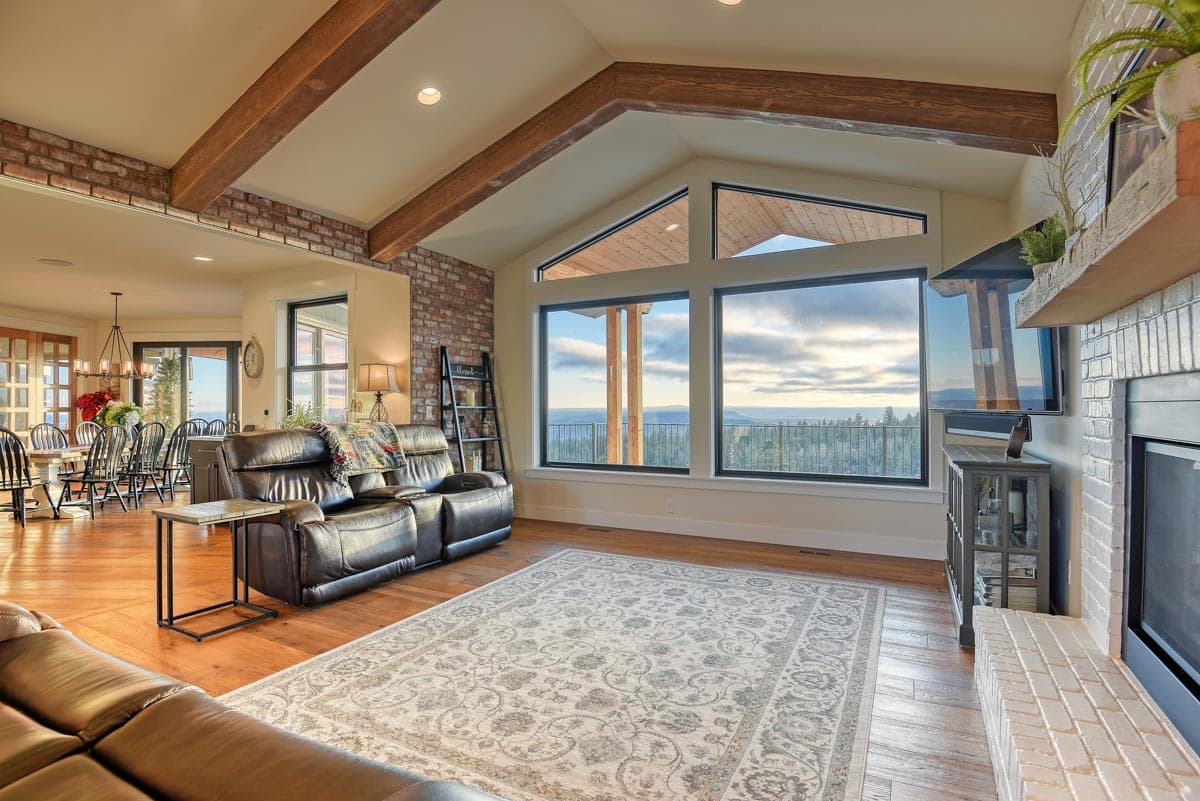 Living room with vaulted ceiling, exposed beams, brick accent walls, and panoramic windows overlooking a landscape.