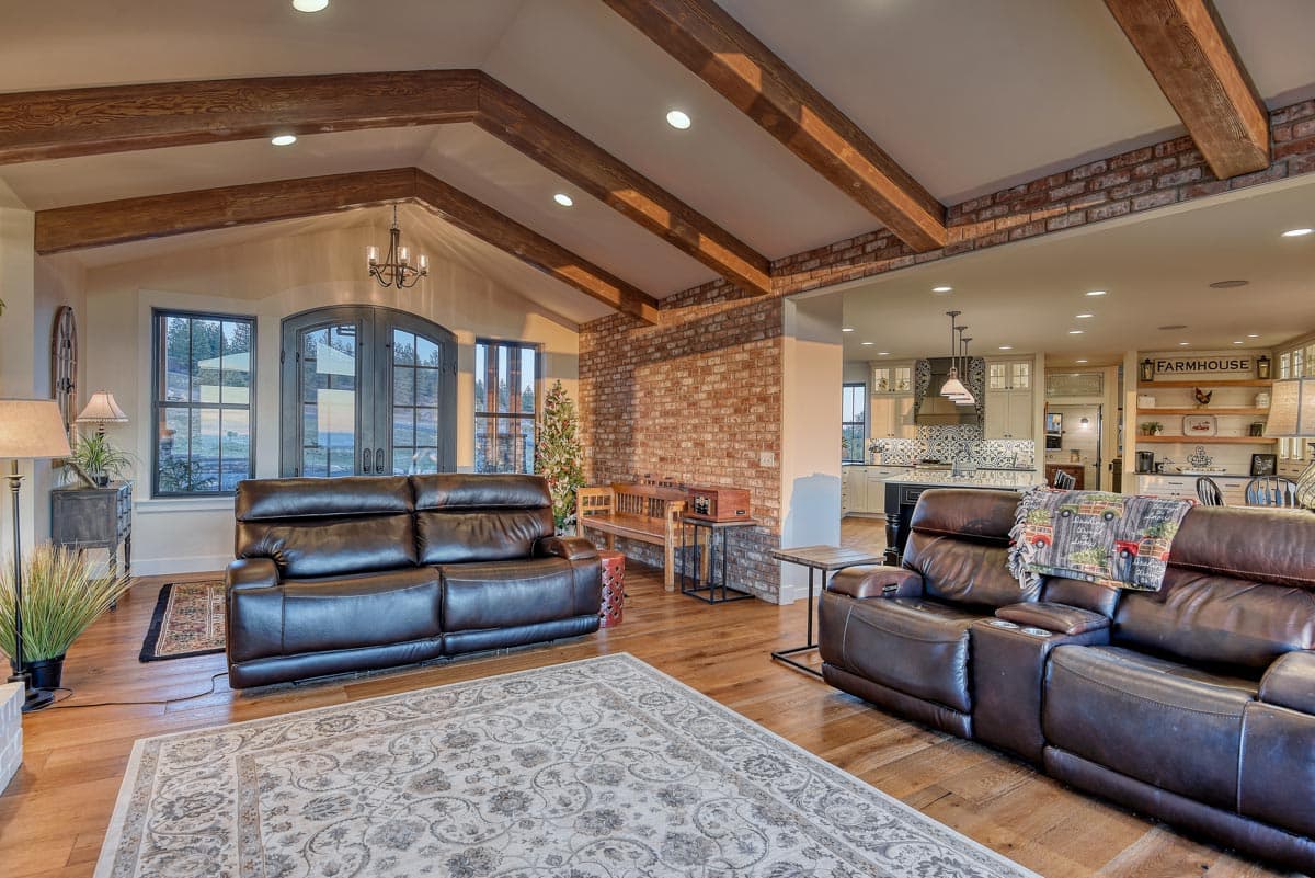 Living room with vaulted ceilings, exposed wood beams, brick accent wall, and open view to kitchen.