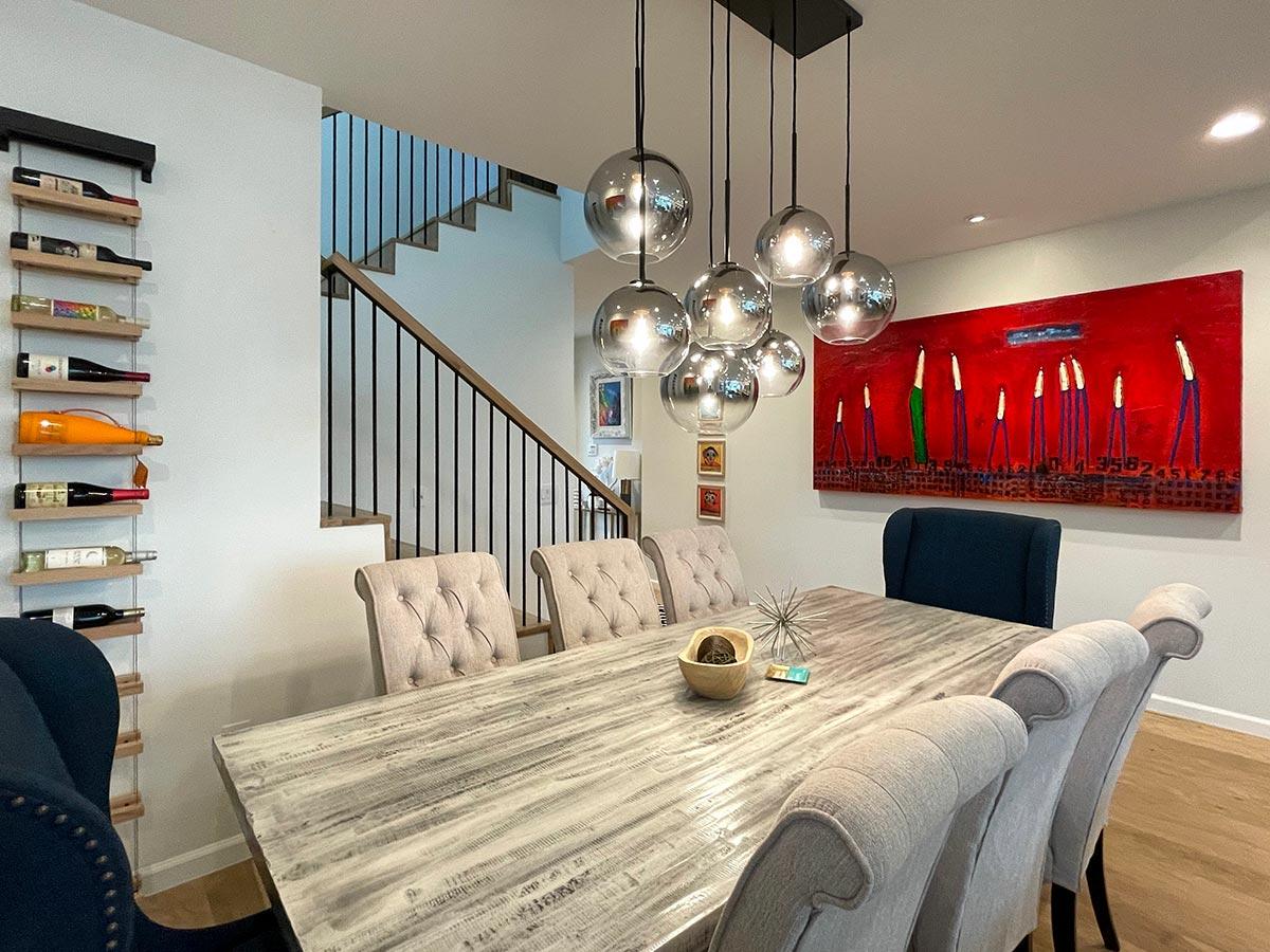Dining room with tufted chairs, modern chandelier, wine rack, and abstract art above a distressed wood table.