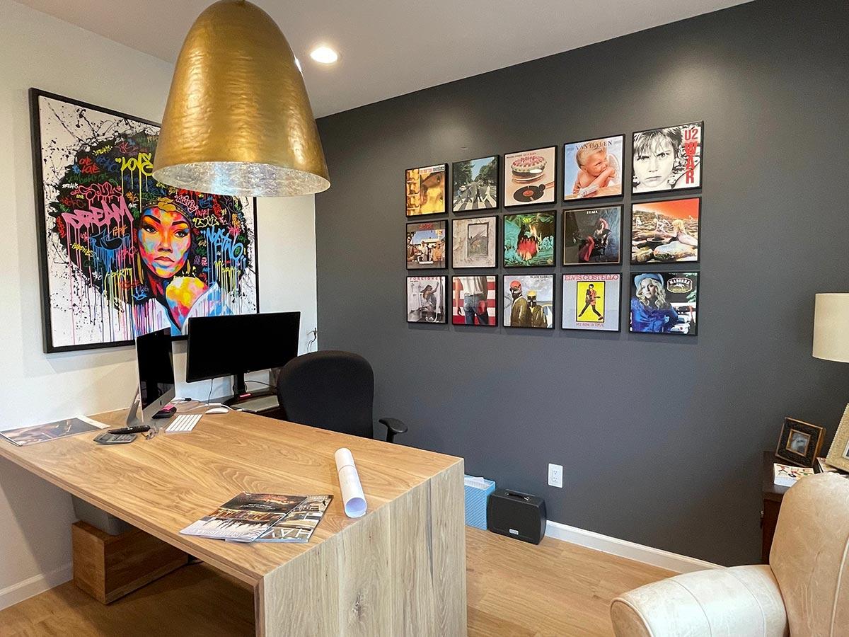 Office interior with a large wooden desk, dual monitors, vibrant wall art, and framed album covers on a dark gray wall.