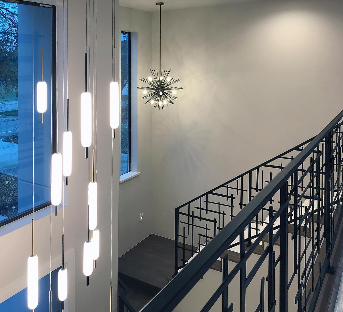 Interior view of a staircase with a modern linear chandelier and a sputnik chandelier, featuring a geometric metal railing.