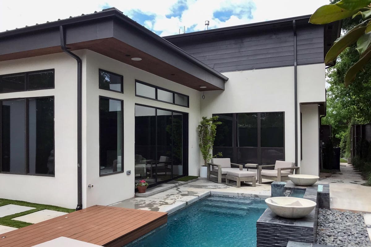 Modern house exterior with stucco finish, dark horizontal siding, flat roof, and expansive dark-framed windows.