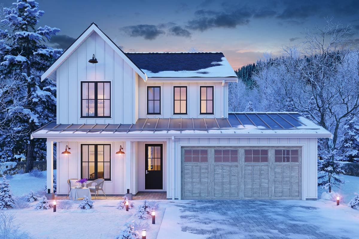 A white farmhouse covered in snow during winter dusk. A front porch with a small table and chairs is lit by lamps. The garage door is closed.