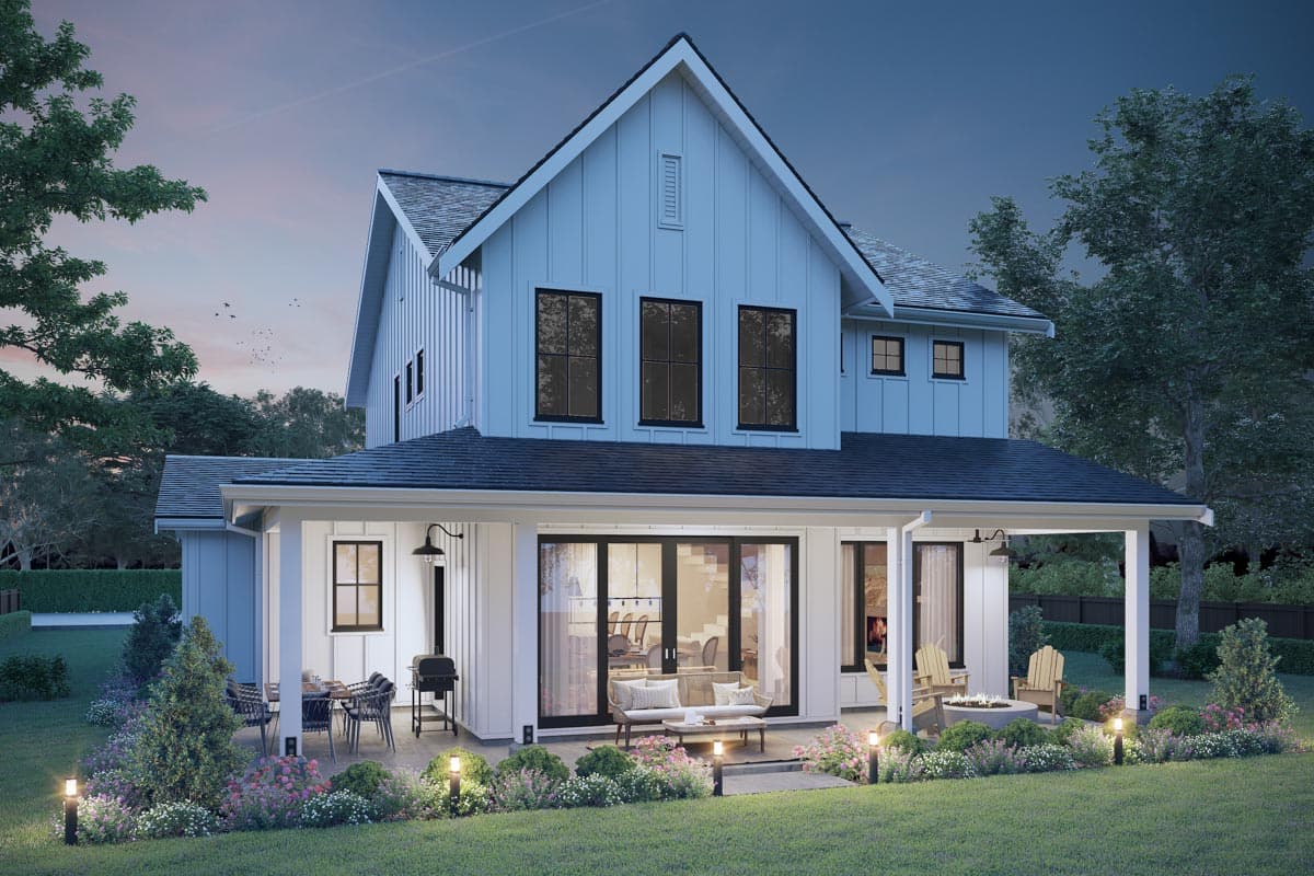 A modern white farmhouse with black trim is lit up at dusk. The back porch features seating, a grill, and a fire pit, with manicured landscaping.