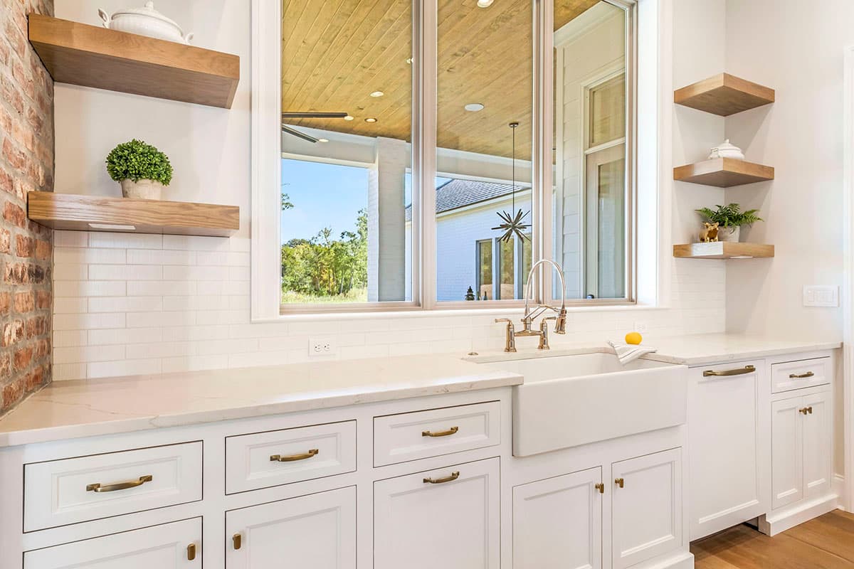 Kitchen interior with white cabinets, farmhouse sink, marble countertop, and open shelving. Includes large windows, and a brick accent wall.
