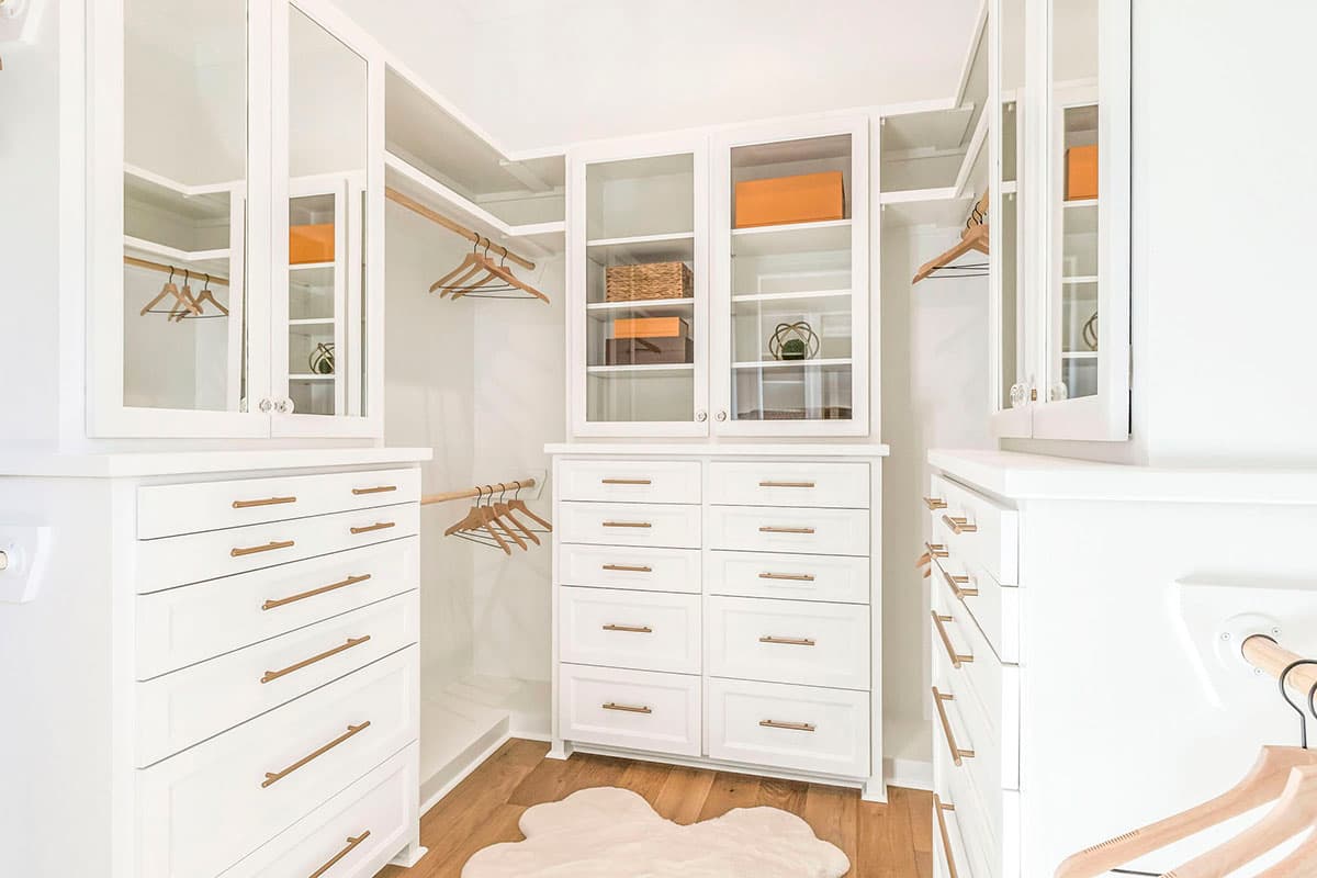 Walk-in closet interior with white cabinetry, glass-front upper cabinets, and a central dresser.  Wooden hanging rods and shelves are visible.