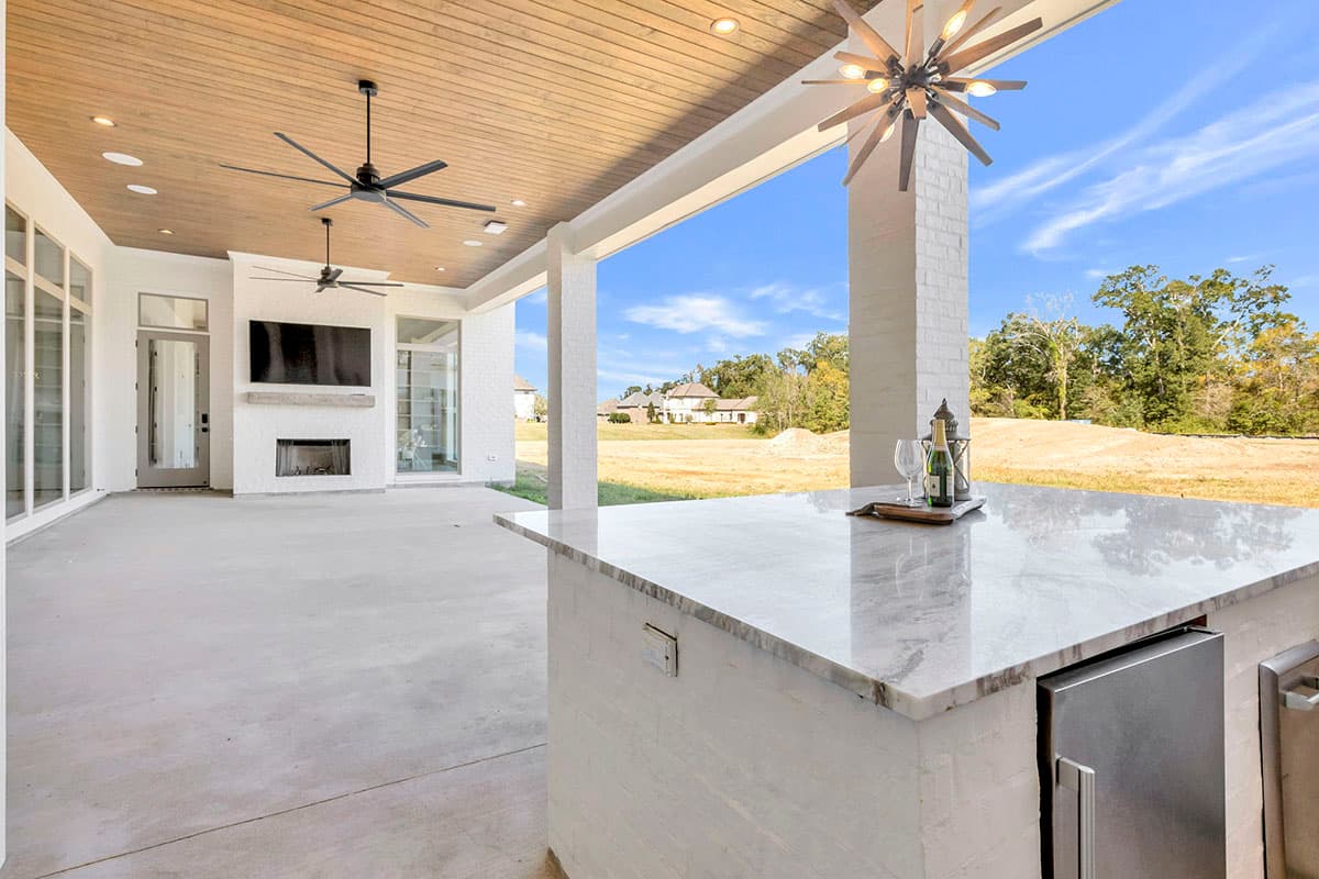 Covered patio with a marble countertop bar, a fireplace with a mounted television, and two ceiling fans. Exposed beam ceiling and brick columns.