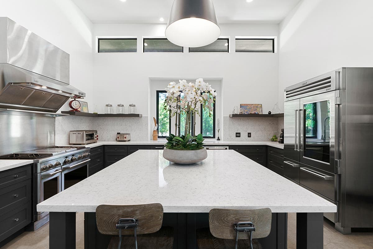 Modern kitchen with a large island, stainless steel appliances, dark cabinetry, and high windows with a white orchid centerpiece.