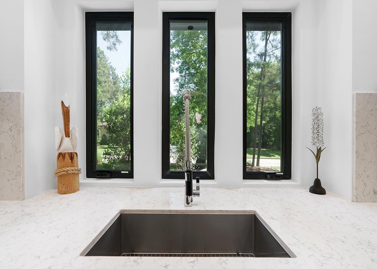 Kitchen view with a large undermount sink, faucet, quartz countertop, and three tall casement windows with black frames.