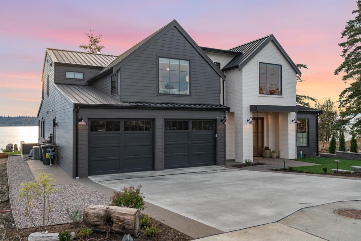Modern farmhouse exterior with white brick and dark siding, featuring a metal roof, gabled dormers, and a three-car garage.
