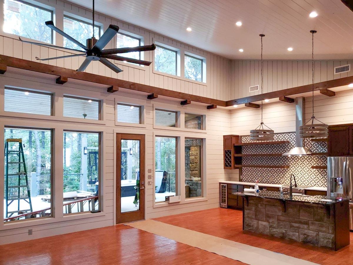 Open concept living space with large windows, exposed beams, wood paneled walls, and a stone-front island in kitchen area.