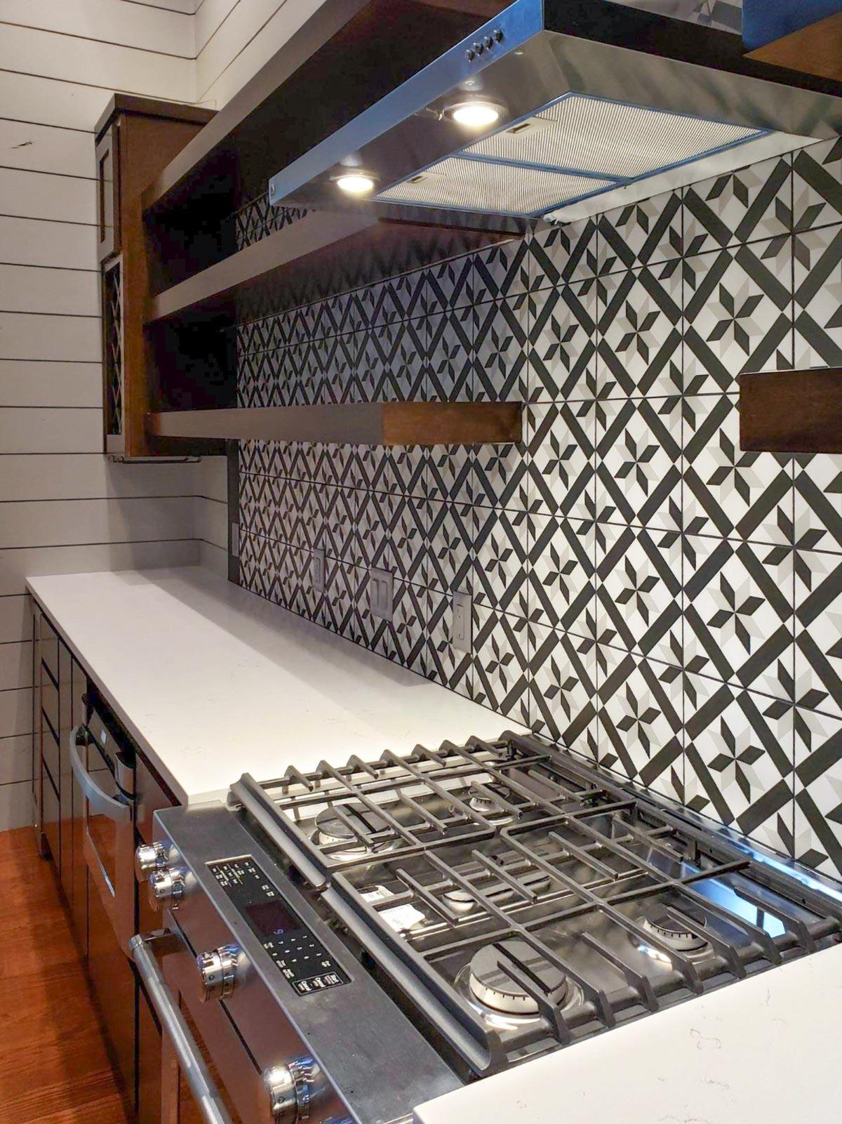 Kitchen backsplash with black and white geometric tile, stainless steel range hood, wooden shelves, and white quartz countertop.
