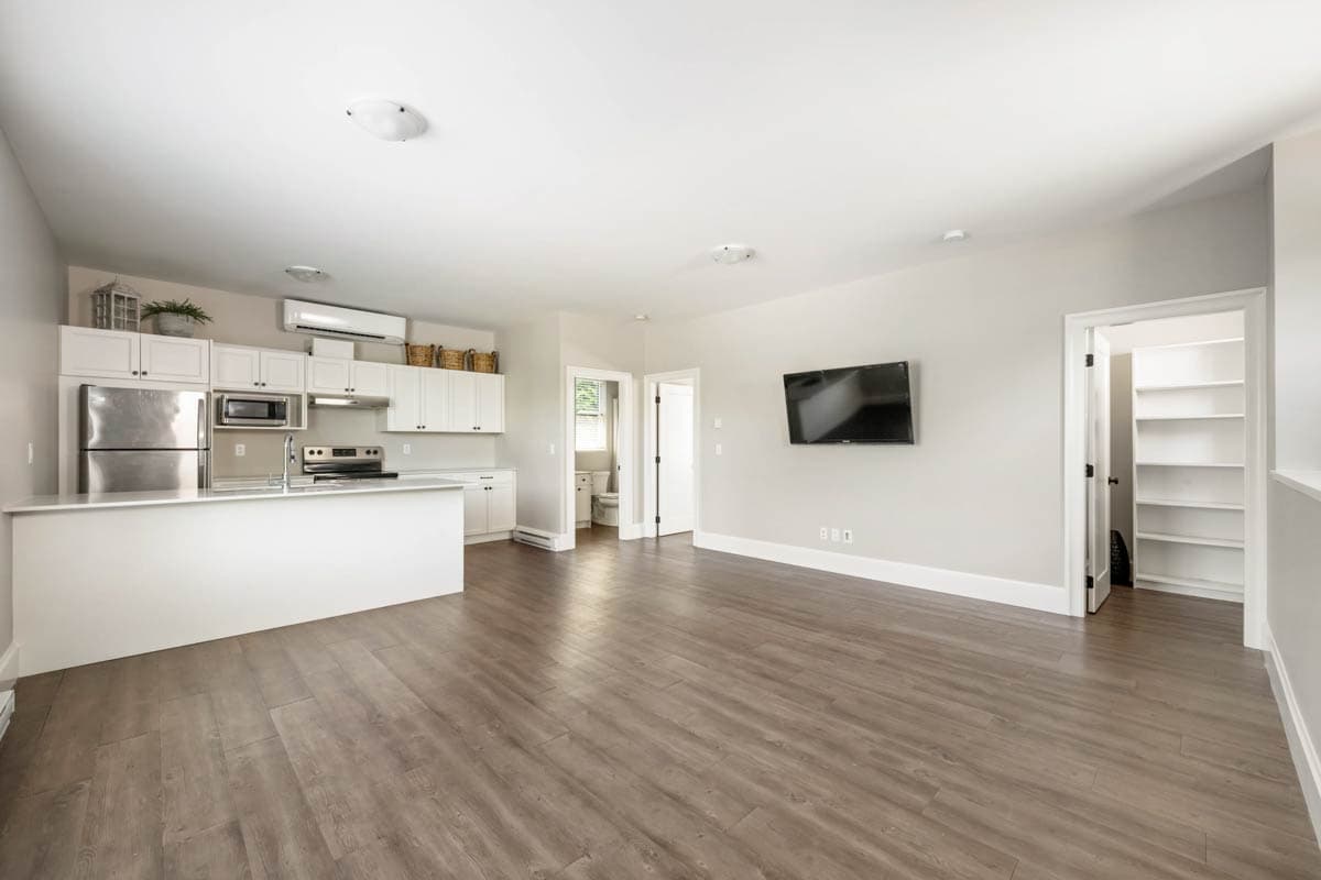 Interior view of a studio apartment with a kitchen island, white cabinets, stainless steel appliances, and a living area with a wall-mounted TV.