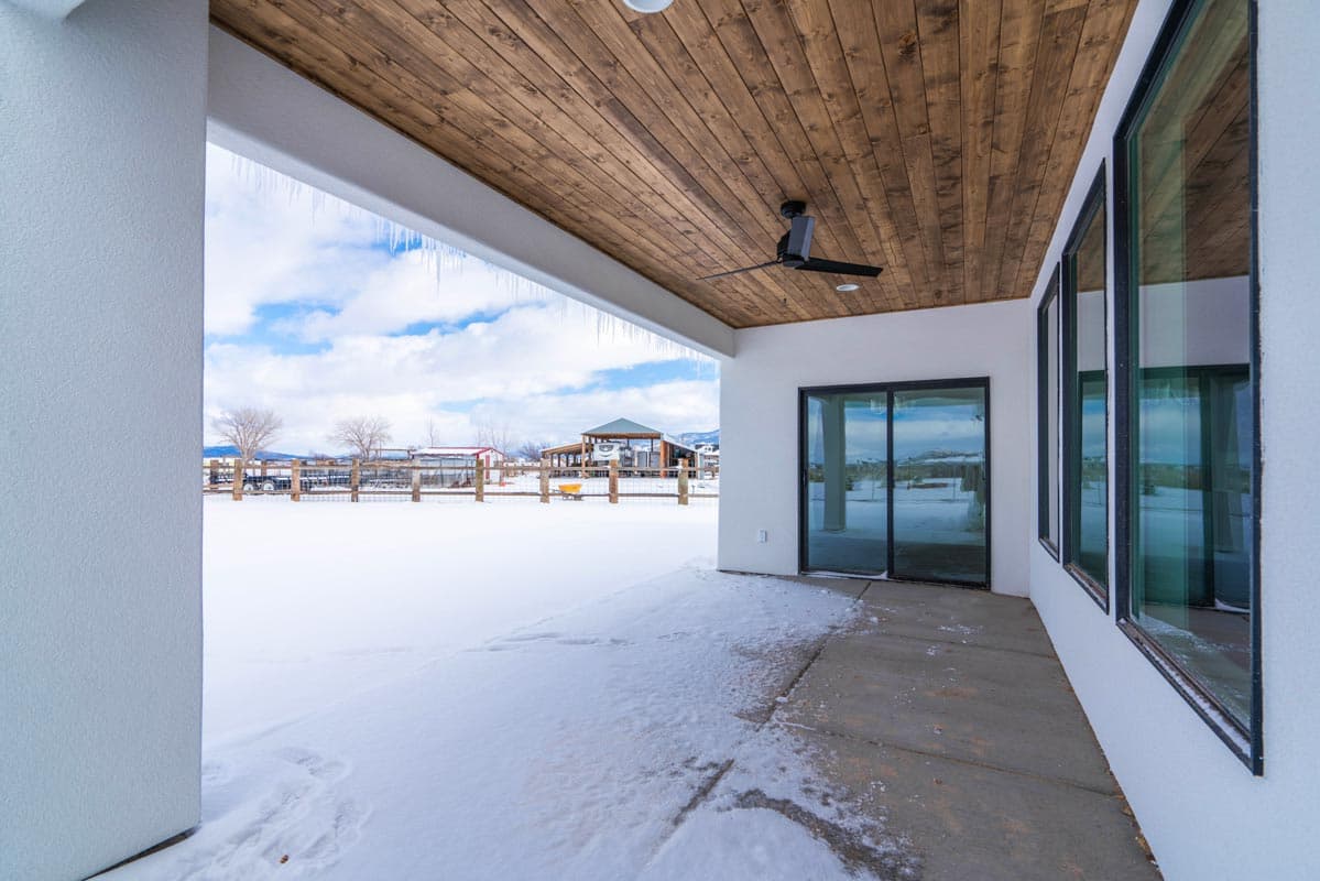 Modern house plan exterior with covered patio, wood ceiling, fan, and large windows overlooking a snowy landscape.