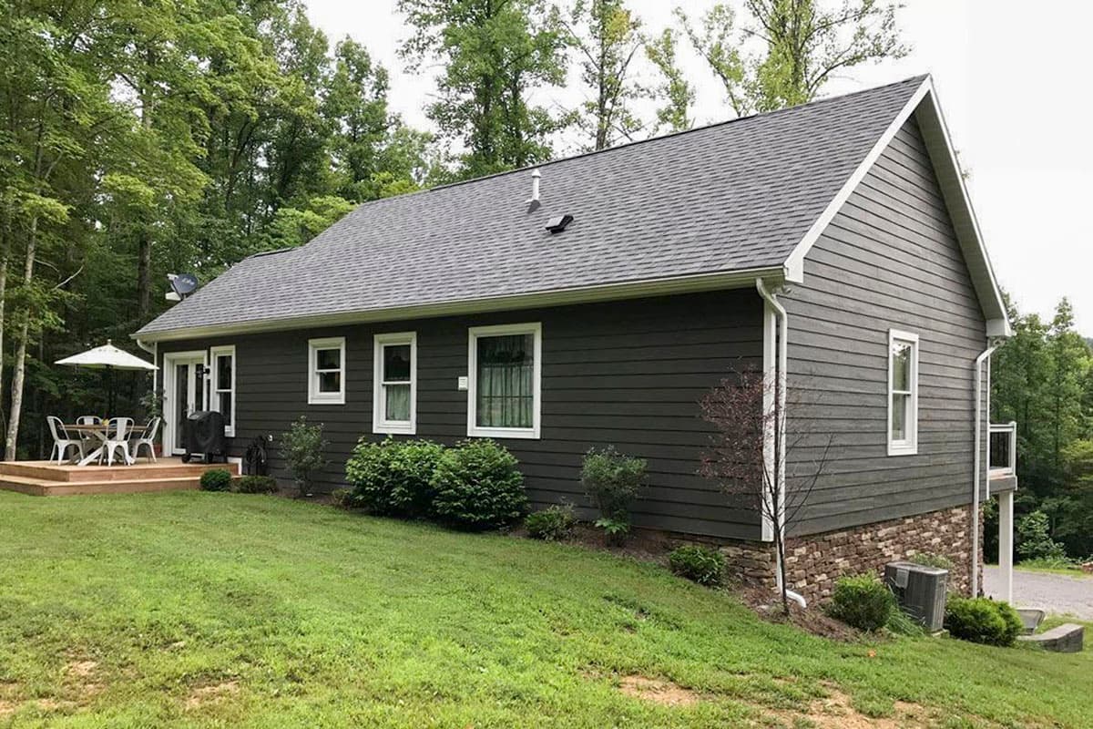 House plan exterior: Ranch-style home with dark siding, stone foundation, prominent gables, and wooden deck with outdoor dining.