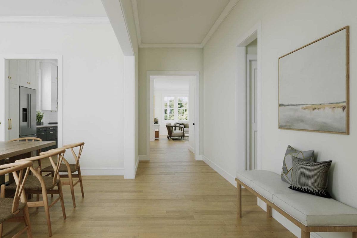 A hallway leads from a dining area with wooden chairs to a living room with an open doorway. A bench with pillows and art hangs on the right wall.
