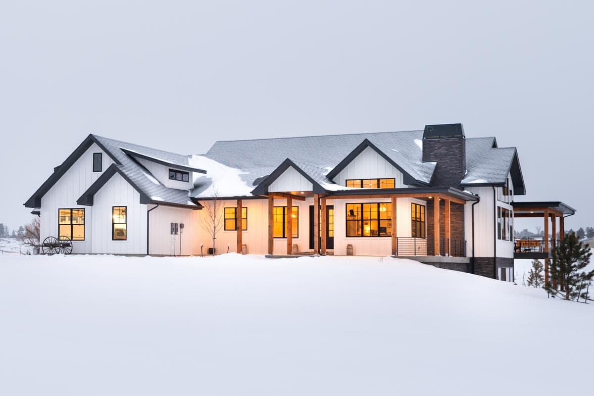 A large white farmhouse with black trim sits in a snowy landscape. The warmly lit windows contrast the cold winter scene and overcast sky.