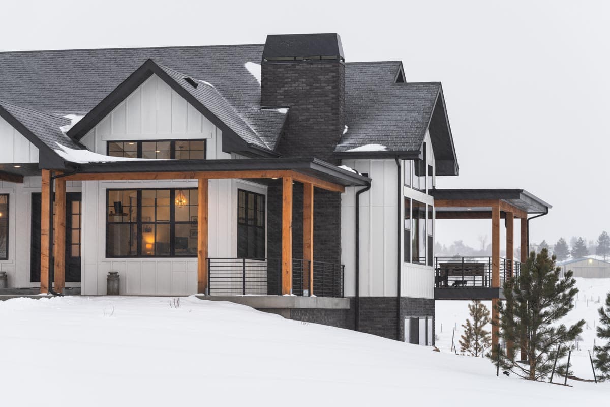 A modern, white farmhouse with black trim and a dark gray roof sits in the snow. Wooden pillars support a covered porch with views of a snowy landscape.