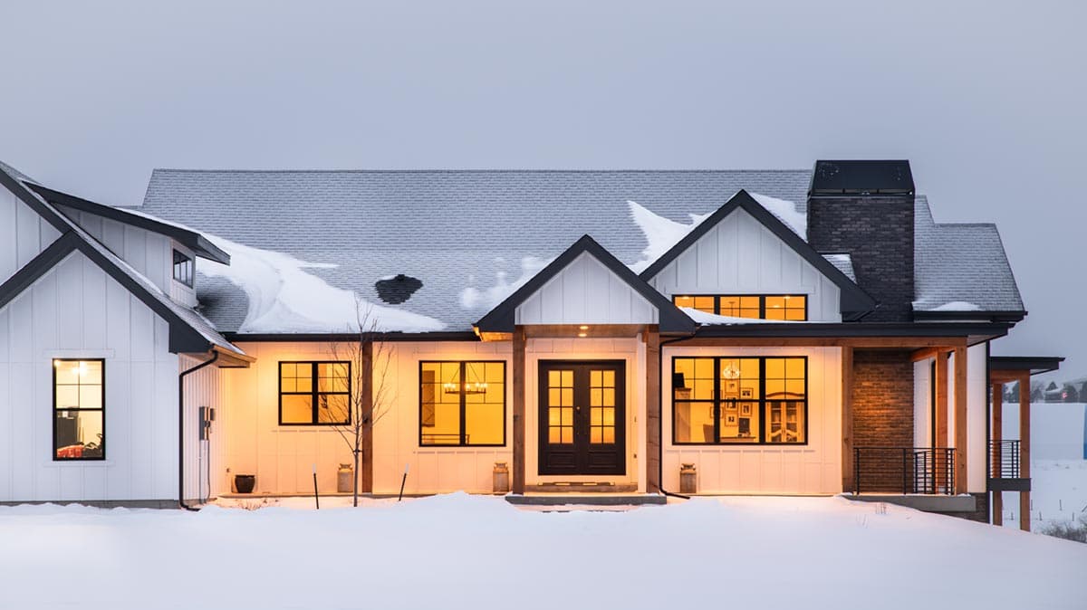 A modern, white farmhouse with black trim and a snow-covered roof at dusk. Warm light glows from windows, inviting contrast with the winter scene.