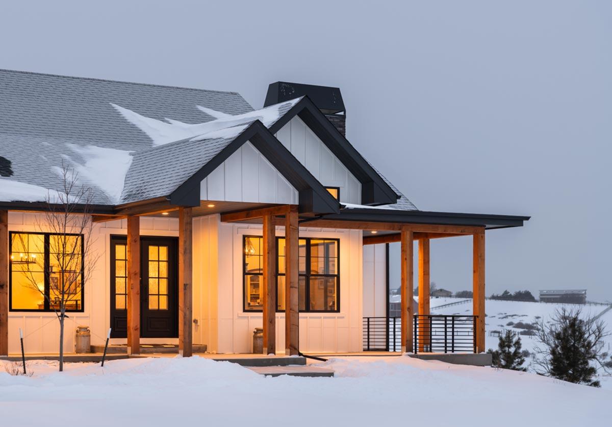 A modern white farmhouse with black trim and a porch, set in a snowy landscape at dusk. The warm interior lights shine through windows and doors.