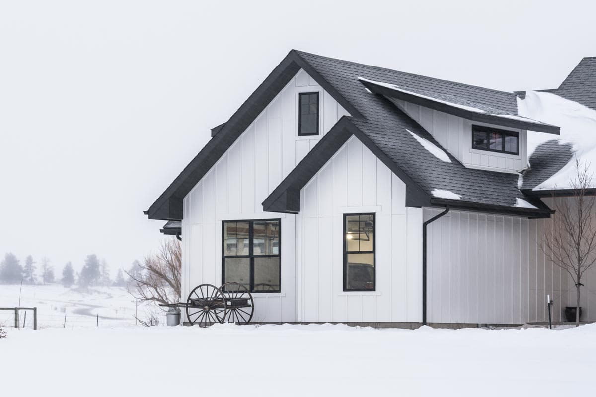 A modern white farmhouse with black trim sits in a snowy landscape. The house has a dark roof and large windows, with a wagon wheel in the foreground.