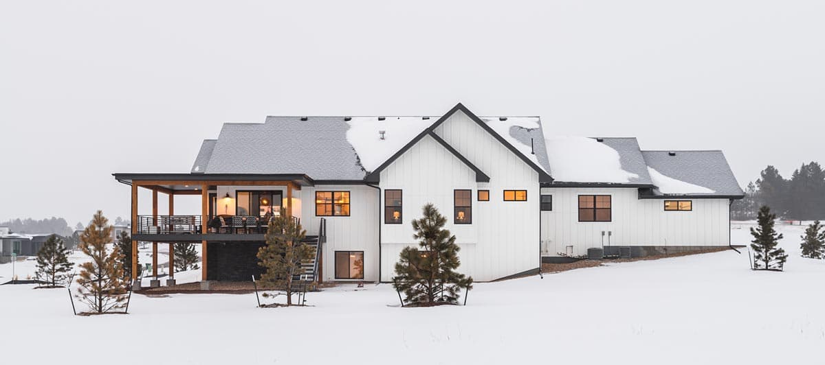 A modern, white farmhouse with a snow-covered roof and surroundings. A covered patio has seating, and the windows glow with warm light.