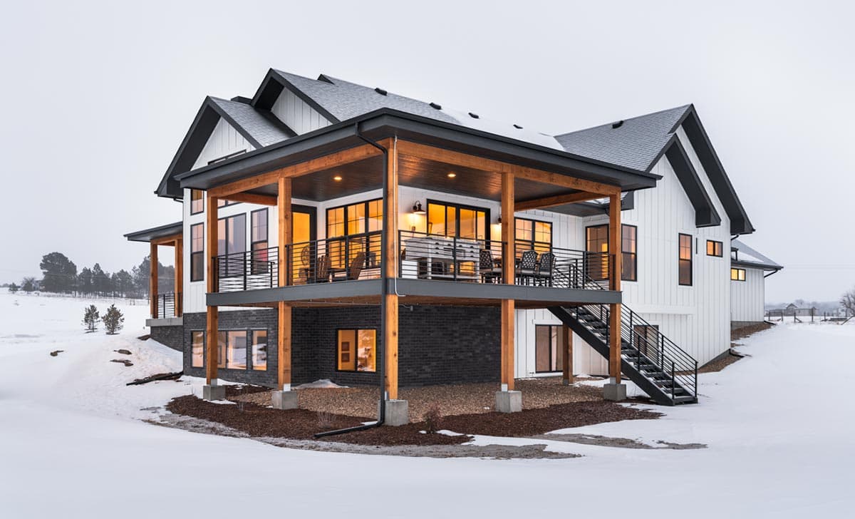 Modern two-story house with a wooden porch and black accents against a snowy landscape. The lights are on inside, suggesting warmth.