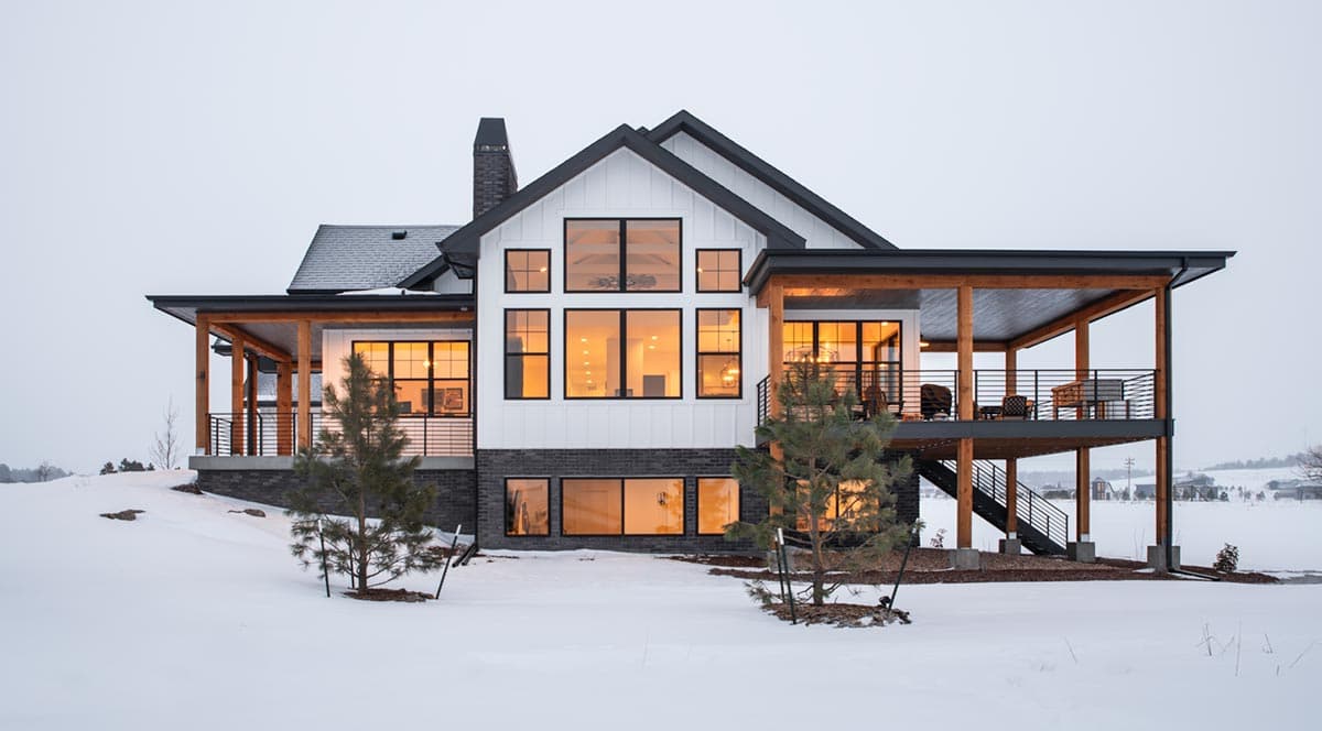 Modern two-story house with large windows and a covered deck, set in a snowy landscape. Exterior features dark trim and a black brick foundation.
