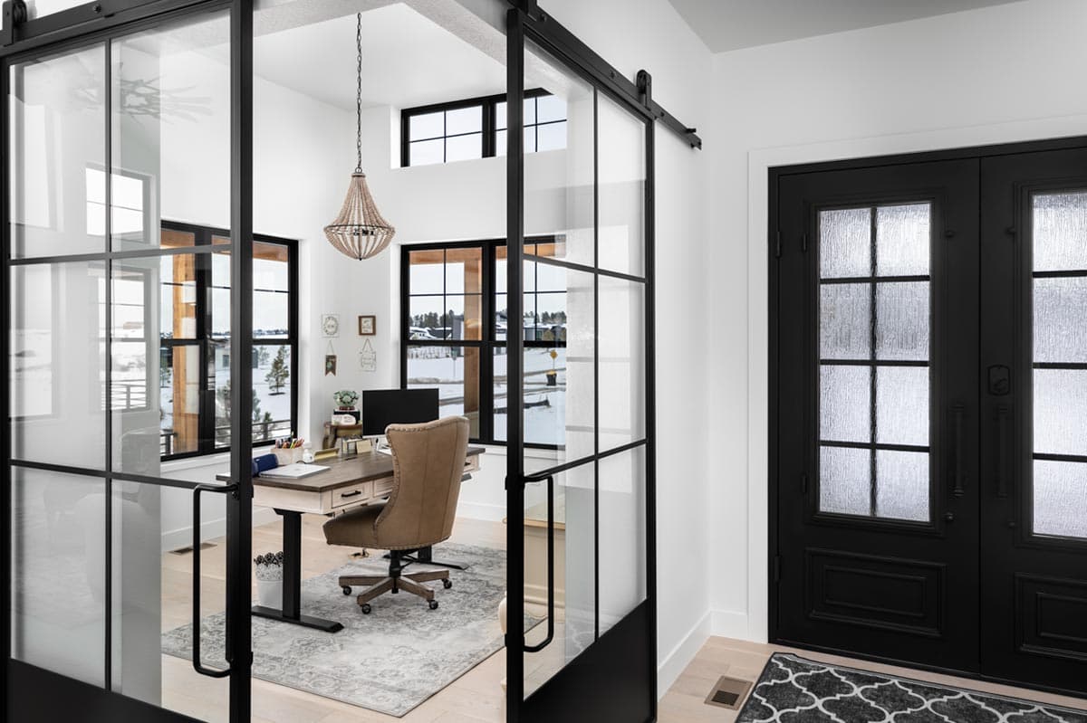 A modern home office with a glass-paneled sliding door, a desk, and a leather chair. Black doors lead to the exterior, and a chandelier hangs above.