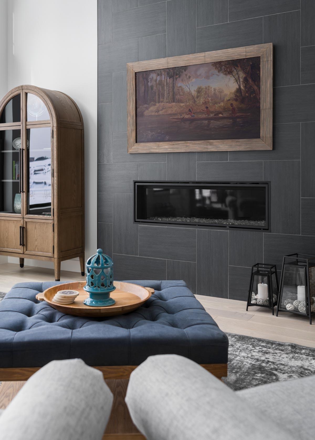 A modern living room with a blue tufted ottoman, fireplace, and artwork. A wooden cabinet stands next to a gray tiled wall with a framed landscape.