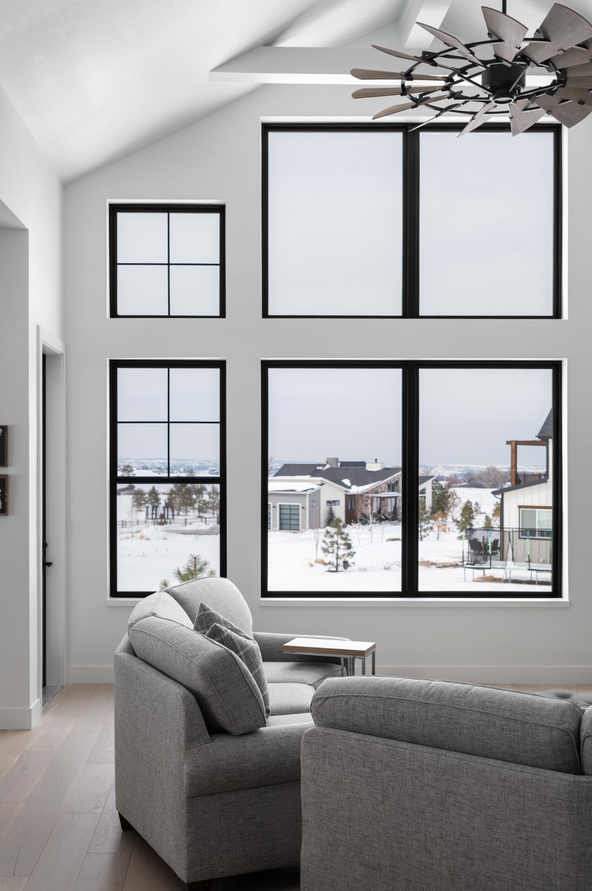 A modern living room featuring multiple large windows with black frames. Through them, a snowy landscape is visible, along with a grey sofa.