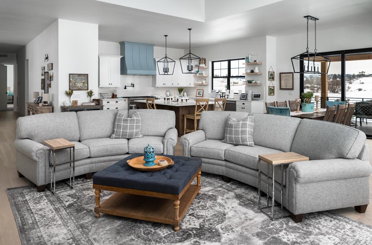 Spacious, open-concept living room with curved gray couches, dark blue ottoman, and a kitchen visible in the background.