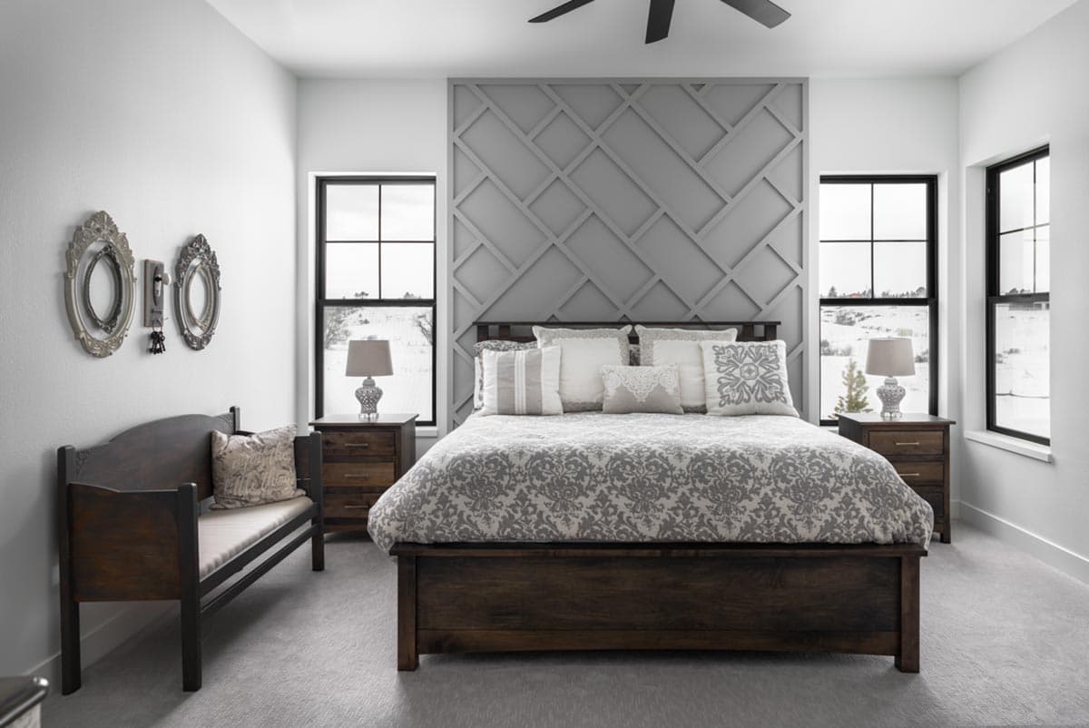 A modern bedroom with a dark wood bed frame and patterned bedding. A decorative wall and two windows flank the bed with matching nightstands on either side.