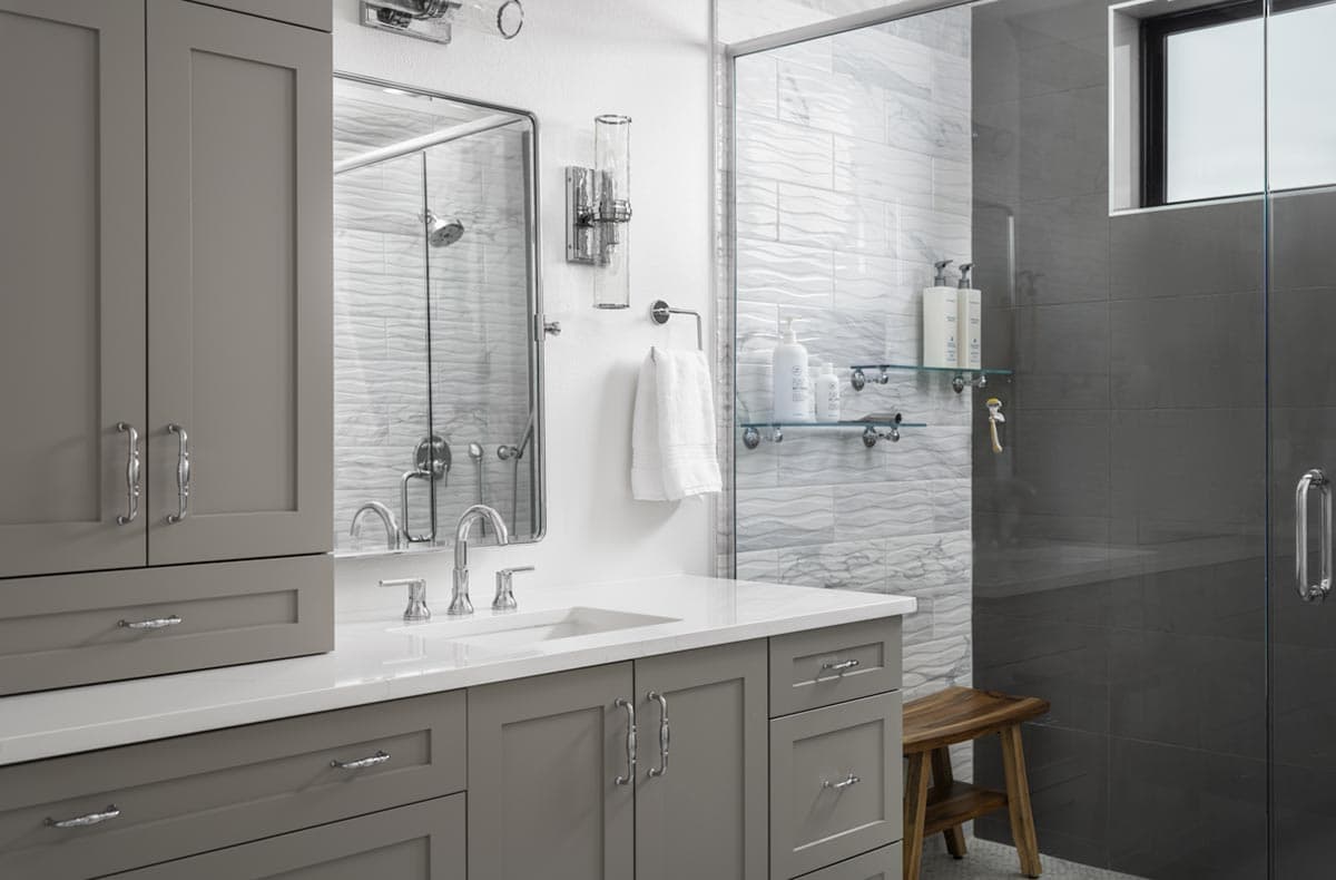 A modern, gray bathroom with a vanity, large mirror, and glass shower. White textured tile provides a backdrop for a wooden stool.