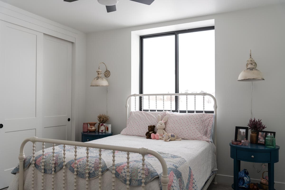 A white iron bed in a child's bedroom with a floral quilt. Soft toys sit on the bed and two vintage lamps flank a large window.