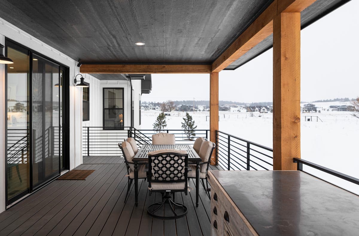 A covered porch with an outdoor dining set overlooks a snowy landscape. The dark-stained deck and black railing contrast the light-colored furniture.