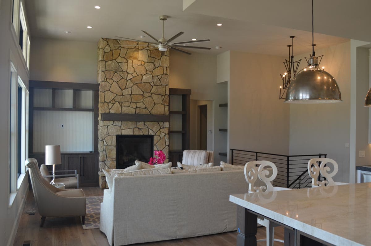 Living room with stone fireplace, built-in shelving, ceiling fan, and large windows.