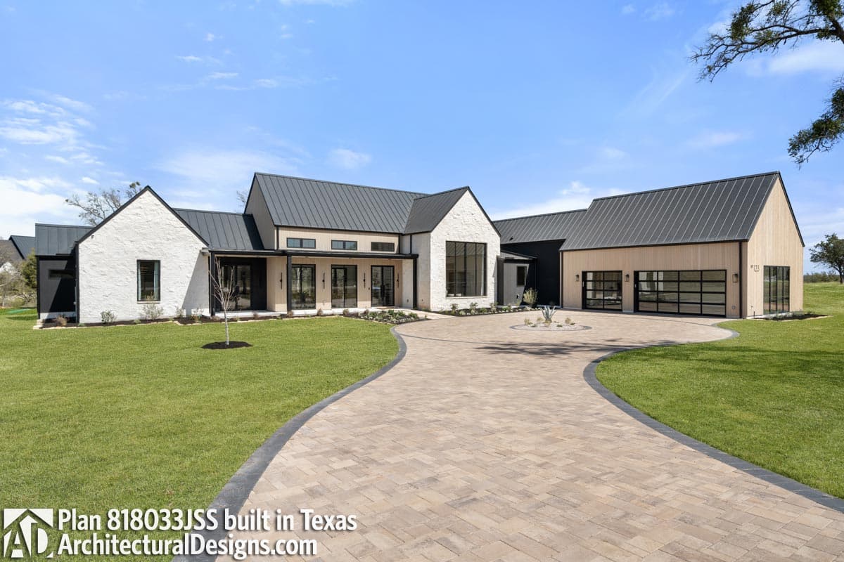 Modern farmhouse with white stucco exterior, black metal roof, and a paved driveway curving to the front. Green lawn surrounds the home.