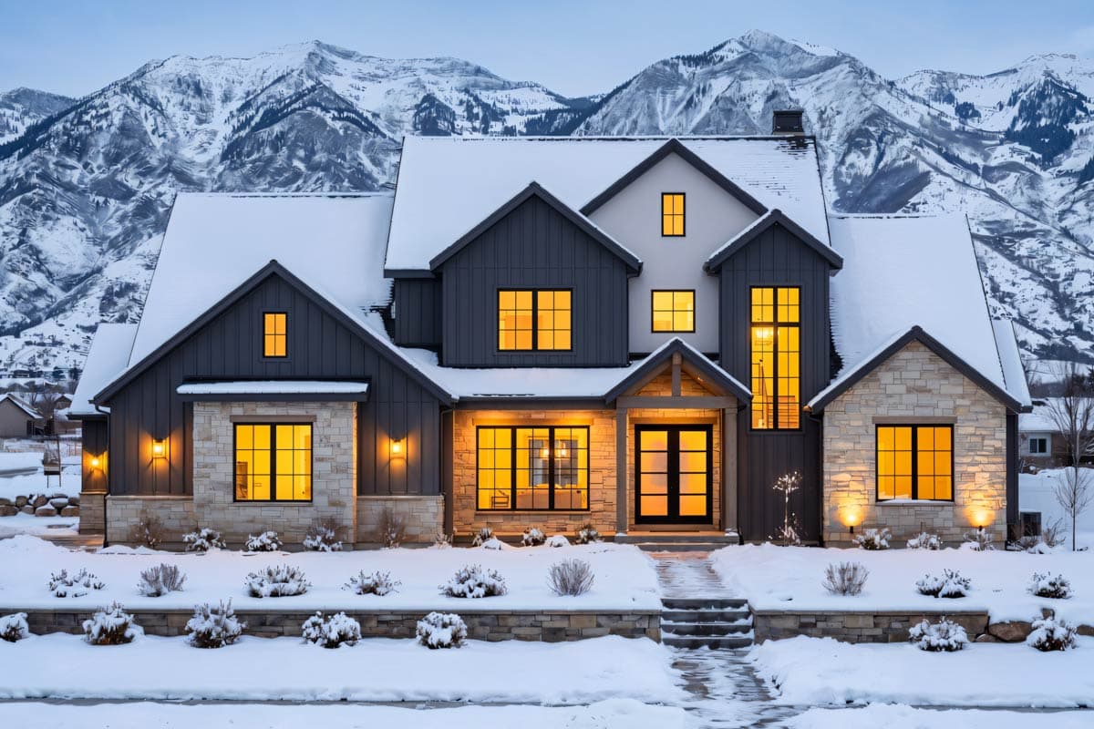 A modern home with lights glowing, nestled beneath snow-covered mountains. Snow blankets the roof, yard, and pathway leading to the front door.