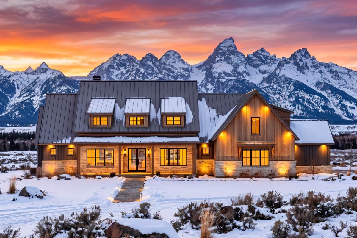 A large, modern home glows with warm light against a snowy landscape and a backdrop of snow-capped mountains at sunset.