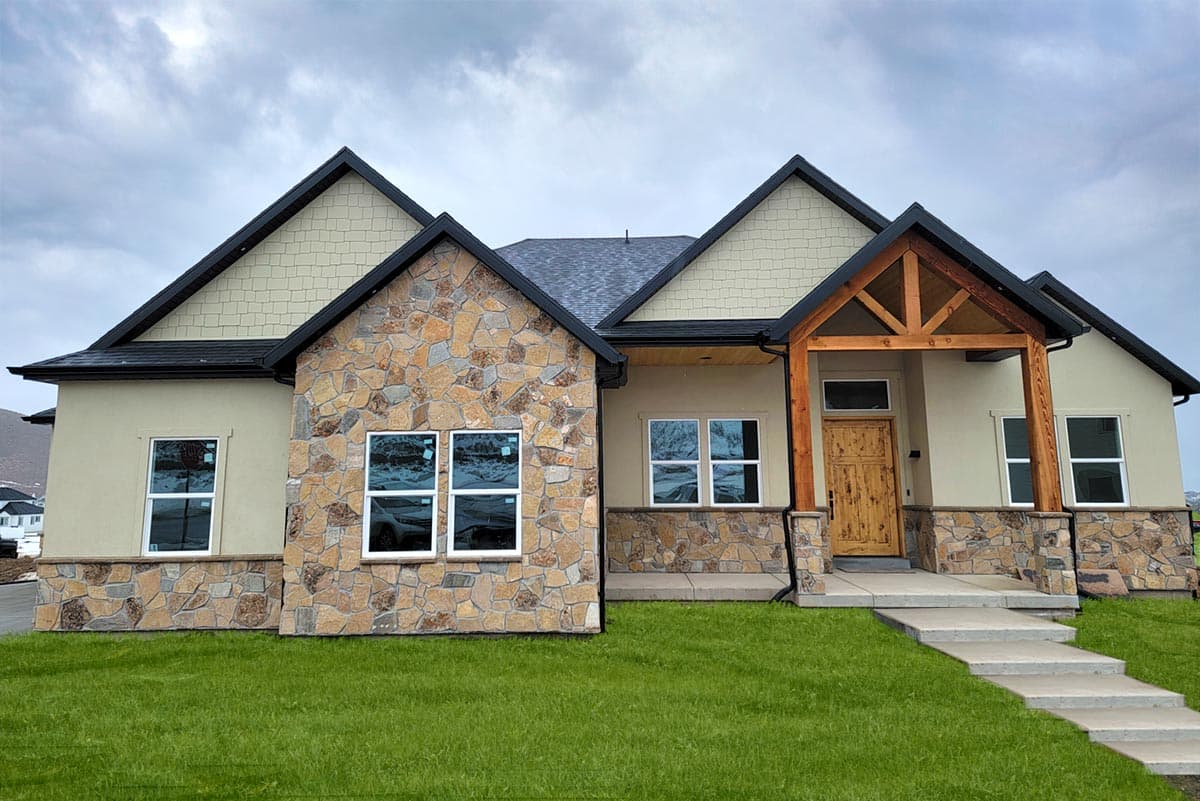 House plan exterior. Ranch style with stone accents and gabled roof. Features include a front porch with exposed timber supports and multiple windows.