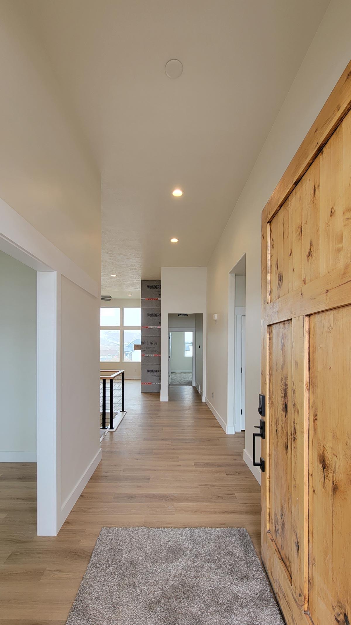 Interior view of a house entryway with a wooden door, hardwood floors, and a hallway leading to rooms with large windows.