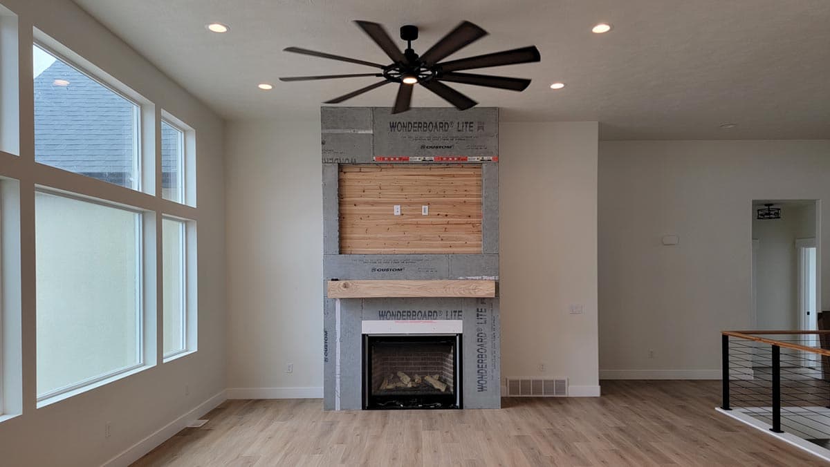 Interior view of a living room with a large fireplace surround, wood accent wall, large windows, and a ceiling fan.